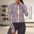 Front view of a woman wearing a vibrant, abstract geometric zip-up athletic hoodie with built-in thumbholes, styled with dark high-waisted leggings in a minimalist fitness studio.