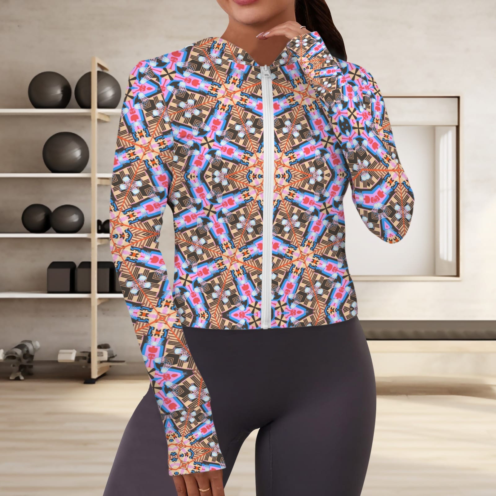 Front view of a woman wearing a vibrant, abstract geometric zip-up athletic hoodie with built-in thumbholes, styled with dark high-waisted leggings in a minimalist fitness studio.