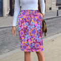 Front view of a woman standing on a city sidewalk wearing a vibrant knee-length drawstring midi skirt featuring neon magenta pink florals styled with a simple white top and brown purse.
