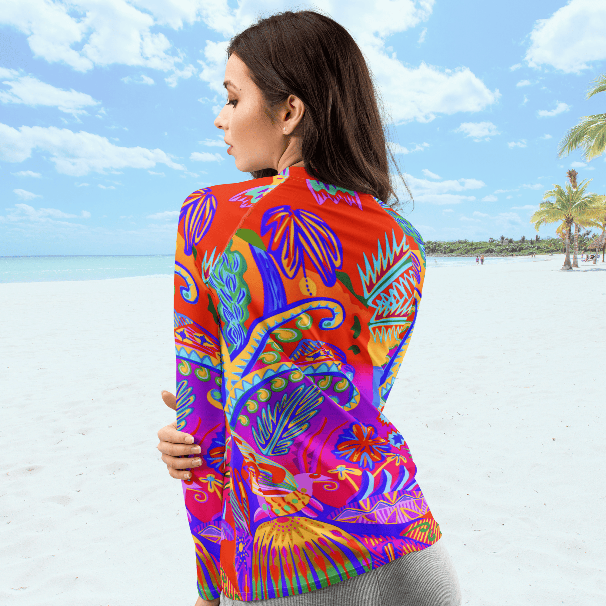 Close up view of female standing on a beach and she is looking to her left. She is wearing a long-sleeved rash guard top with a vibrant, colorful tropical scene with insects. The top features a red base with blue, orange, purple, and yellow floral and abstract patterns.