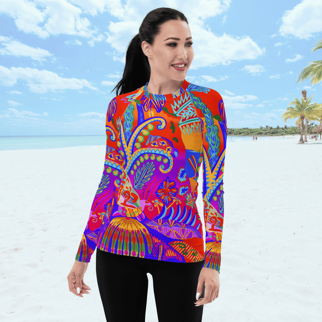 Front view of a woman with black hair tied up in black ponytail smiles while wearing a vibrant, long-sleeved rash guard. The top features an intricate, multicolored tropical print with plants, trees and insects. She is standing on a white sand beach under a bright blue sky.