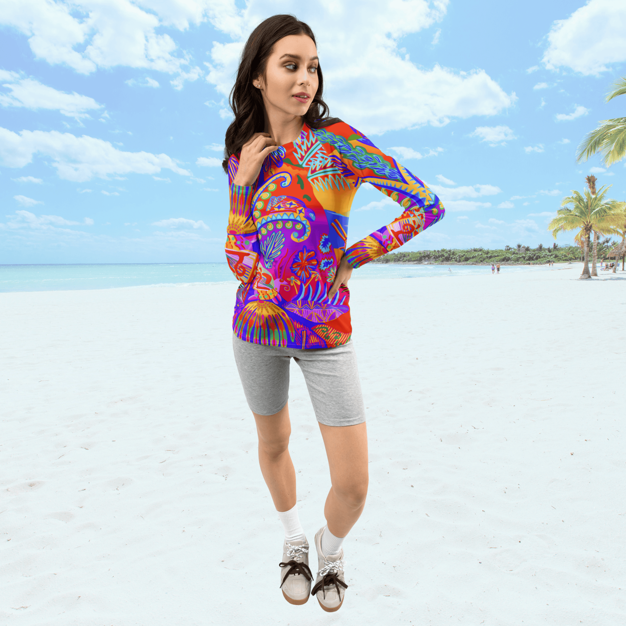Full front view of female standing on a beach. She is wearing a a long-sleeved rash guard top with a vibrant, colorful tropical scene with insects. The top features a red base with blue, orange, purple, and yellow floral and abstract patterns.