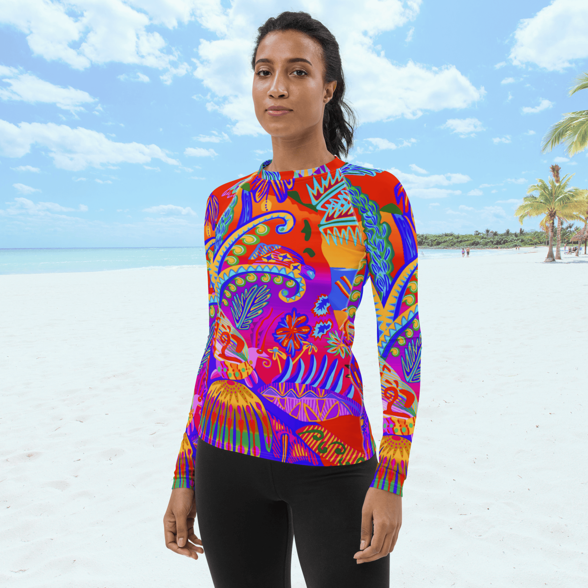 Front view of a woman with black hair tied up in black ponytail wearing a vibrant, long-sleeved rash guard. The top features an intricate, multicolored tropical print with plants, trees and insects. She is standing on a white sand beach under a bright blue sky.