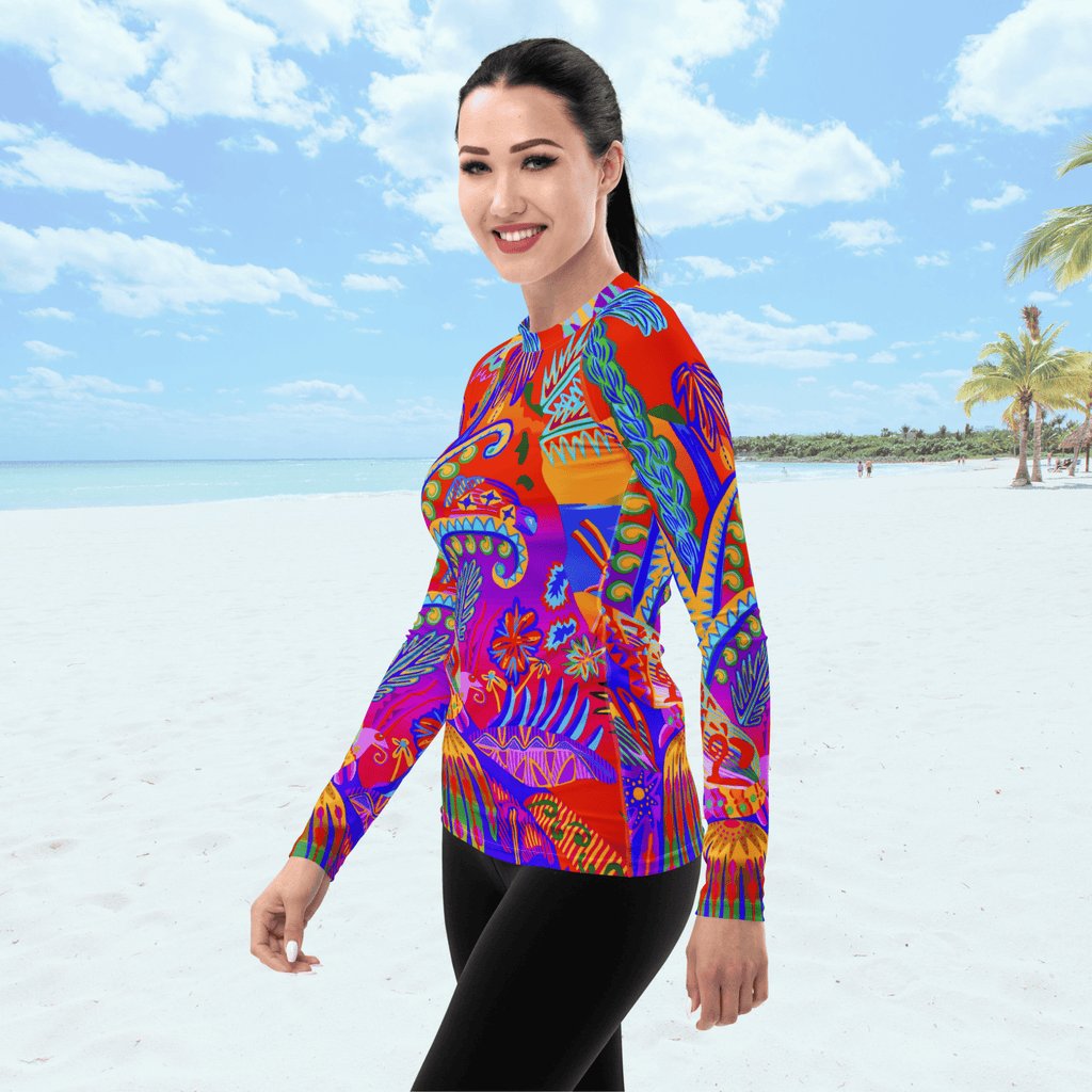 Angle front view of female standing on a beach. She is wearing a a long-sleeved rash guard top with a vibrant, colorful tropical scene with insects. The top features a red base with blue, orange, purple, and yellow floral and abstract patterns.