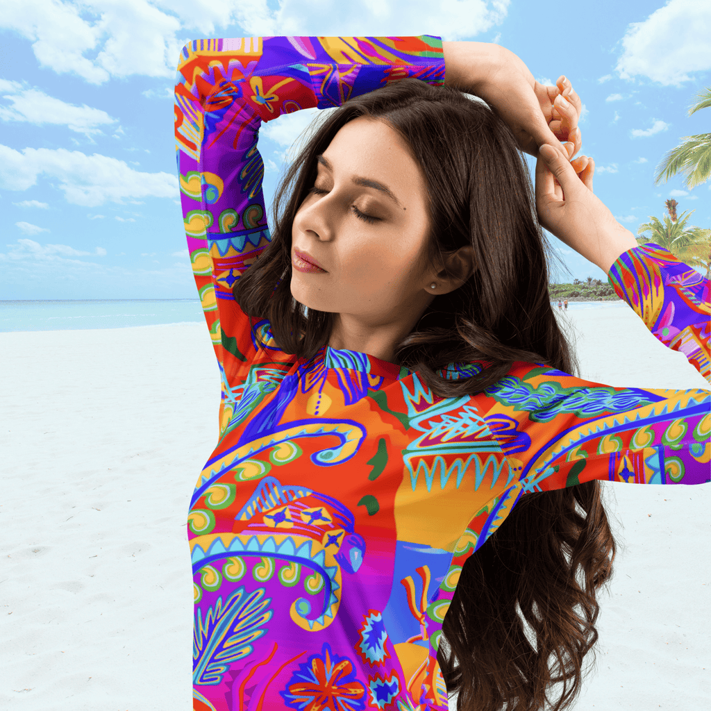 Close up angle front view of female standing on a beach. She is wearing a a long-sleeved rash guard top with a vibrant, colorful tropical scene with insects. The top features a red base with blue, orange, purple, and yellow floral and abstract patterns.