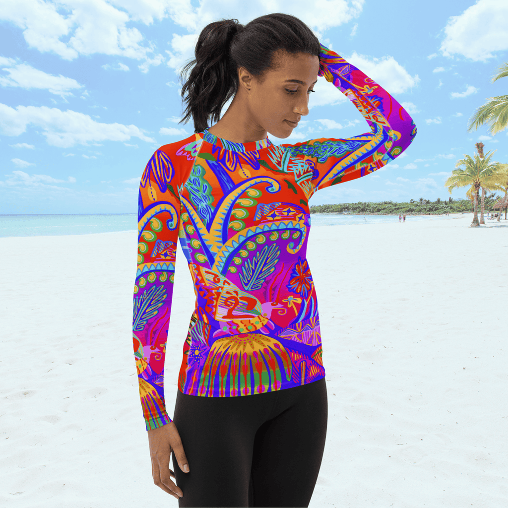 Angle front view of female standing on a beach. She is wearing a a long-sleeved rash guard top with a vibrant, colorful tropical scene with insects. The top features a red base with blue, orange, purple, and yellow floral and abstract patterns. She is wearing a a long-sleeved rash guard top with a vibrant, colorful tropical scene with insects. The top features a red base with blue, orange, purple, and yellow floral and abstract patterns.