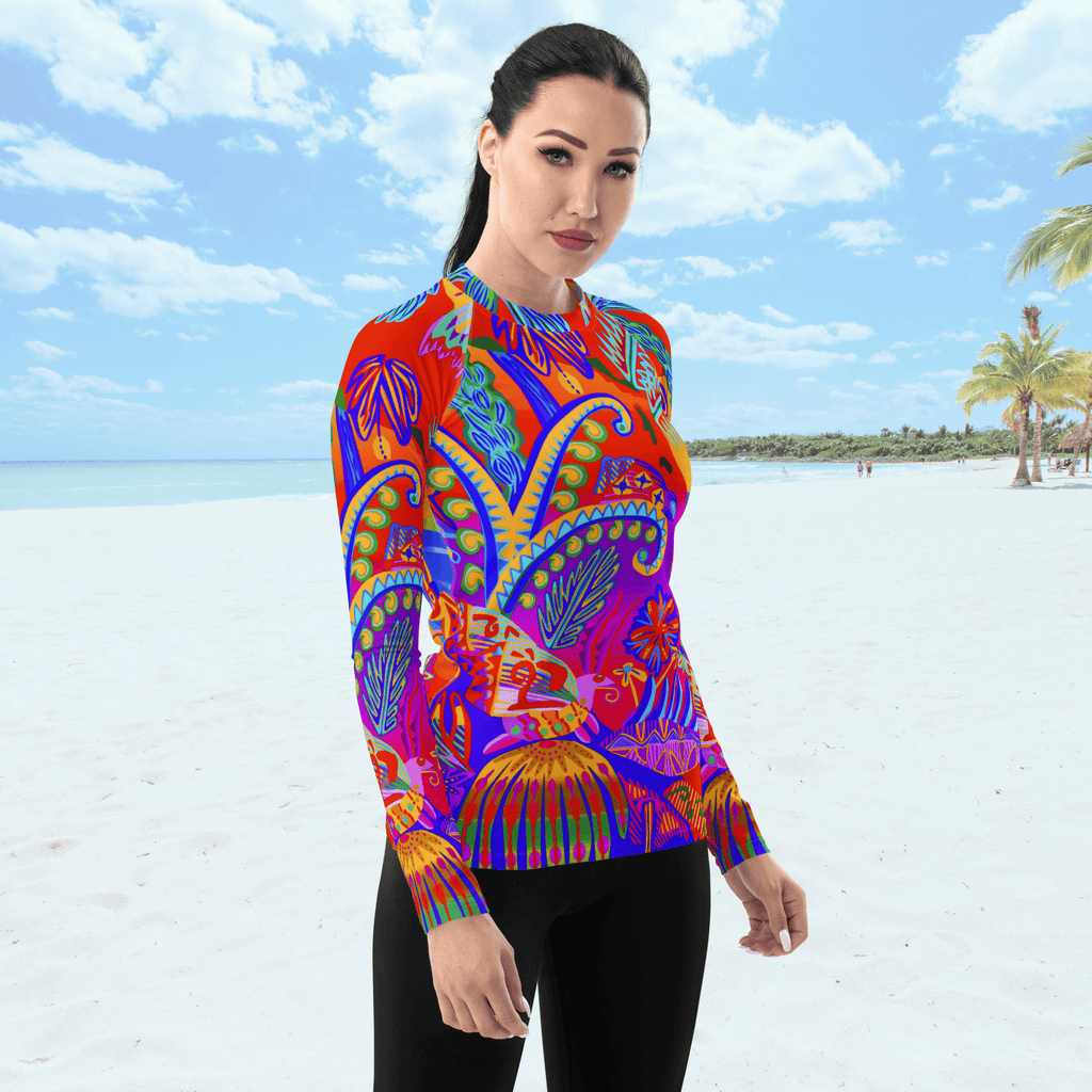Angle front view of female standing on a beach. She is wearing a a long-sleeved rash guard top with a vibrant, colorful tropical scene with insects. The top features a red base with blue, orange, purple, and yellow floral and abstract patterns.