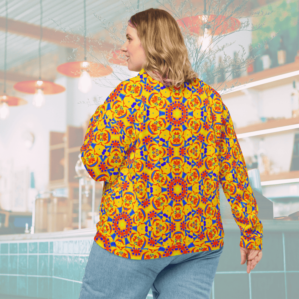 Rear three-quarter view of a blonde woman wearing a vibrant yellow bomber jacket with an all-over orange, red, and blue geometric mandala pattern, paired with light-wash denim jeans in a blurred cafe setting.