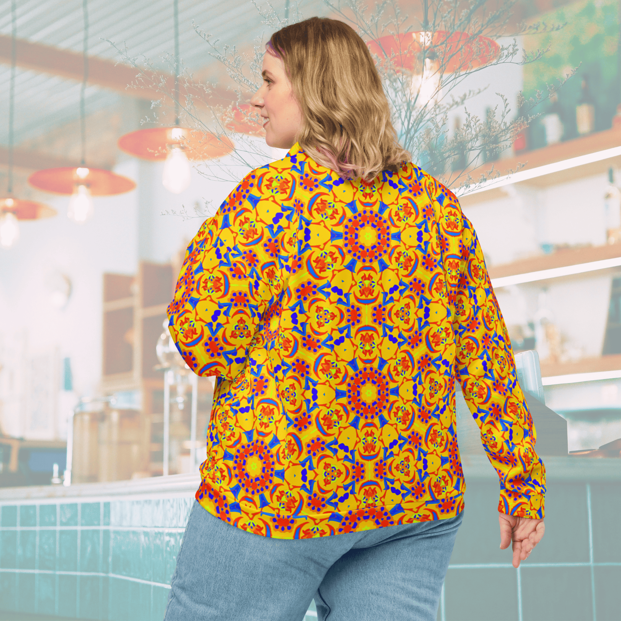 Rear three-quarter view of a blonde woman wearing a vibrant yellow bomber jacket with an all-over orange, red, and blue geometric mandala pattern, paired with light-wash denim jeans in a blurred cafe setting.