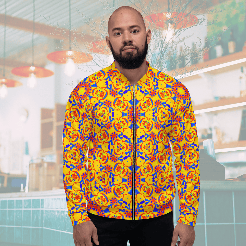 Front view of a bearded man wearing a yellow zip-up bomber jacket showcasing a bold blue, red, and orange kaleidoscope mandala design, styled with black jeans against a cafe interior backdrop.