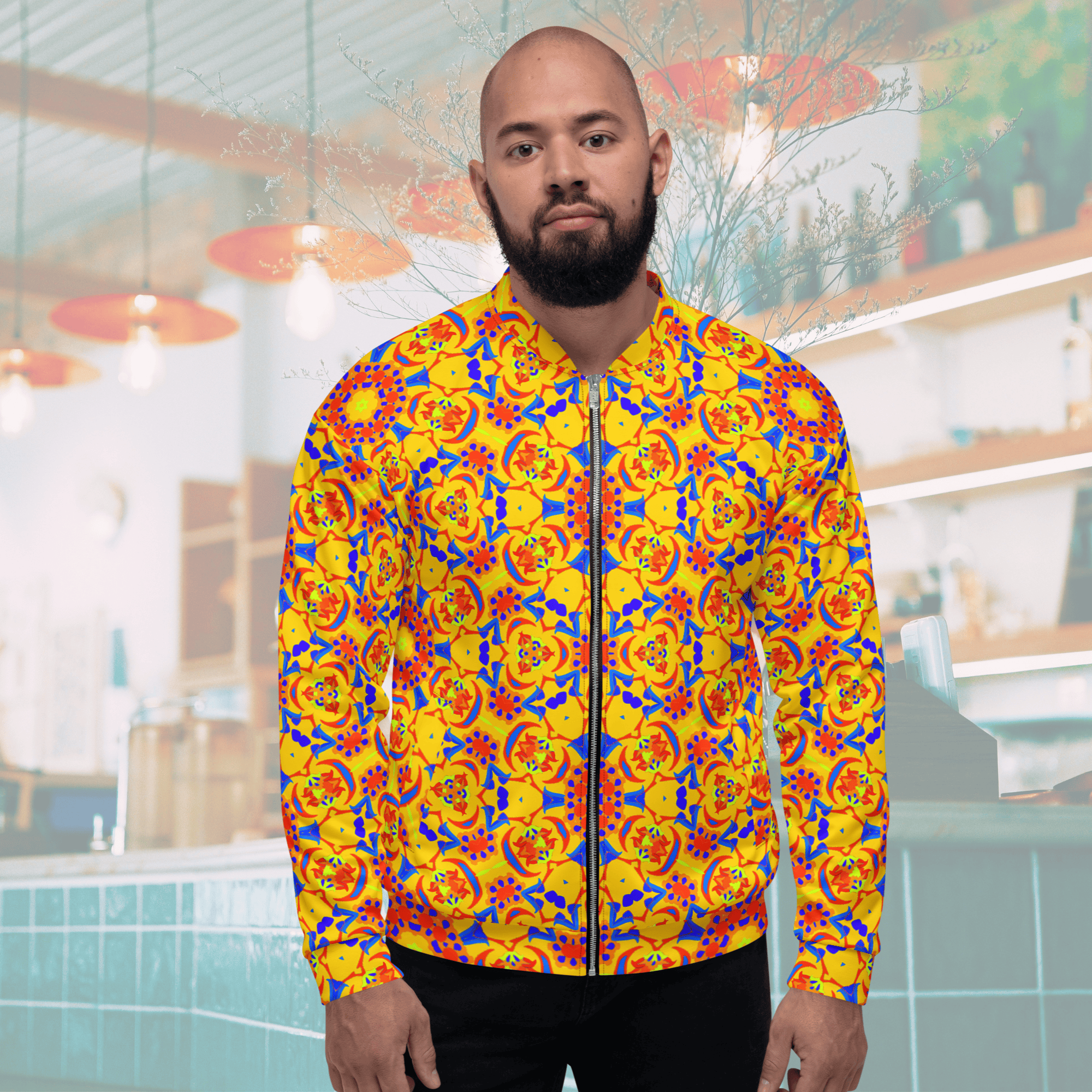 Front view of a bearded man wearing a yellow zip-up bomber jacket showcasing a bold blue, red, and orange kaleidoscope mandala design, styled with black jeans against a cafe interior backdrop.