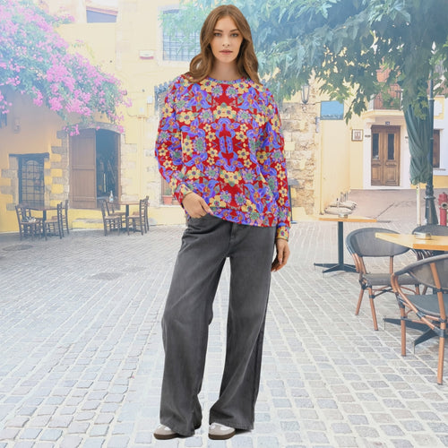 Front view of a woman wearing a vivid red long-sleeve top with a bold floral and abstract pattern in purple, blue, yellow, and pink, standing in a sunny outdoor street café setting.
