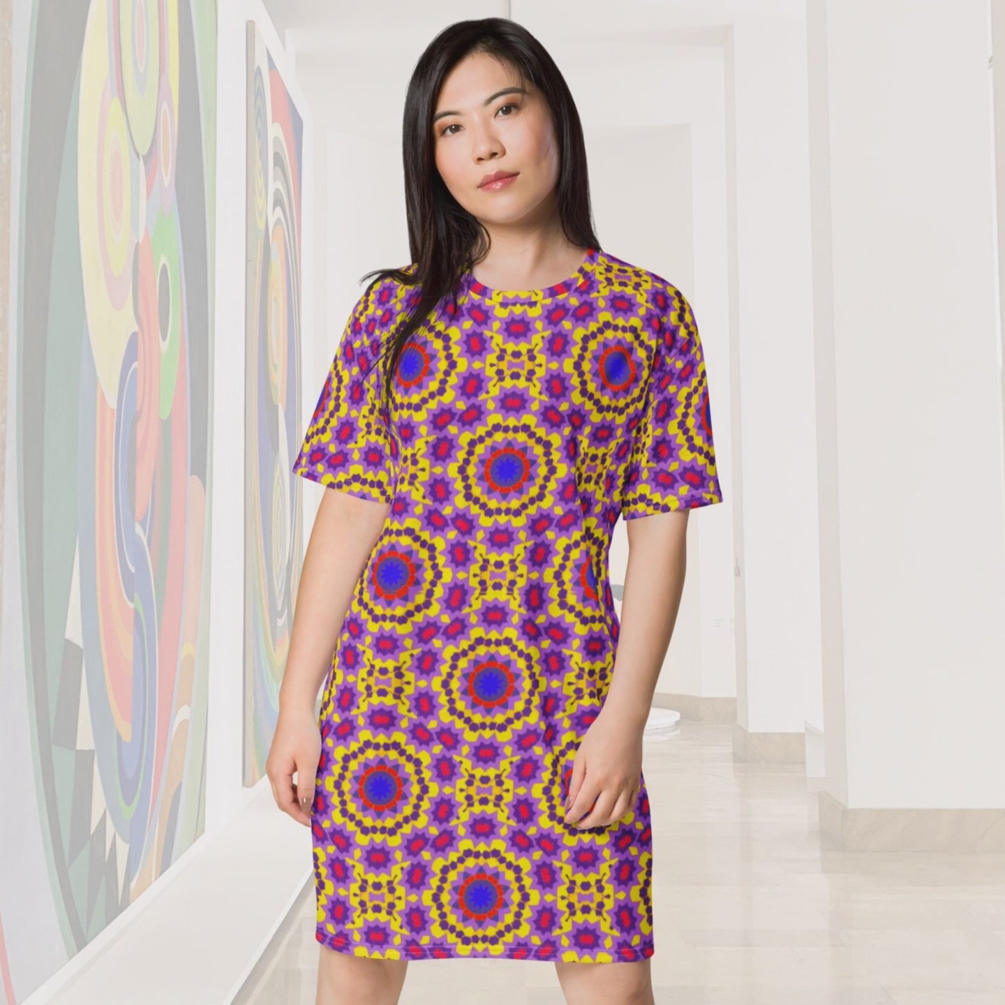 A 10-second video of a woman with long dark hair walking confidently toward the camera through a bright, minimalist art gallery. She is wearing a short-sleeved, knee-length dress featuring a vibrant, repeating geometric pattern of purple, yellow, and red mandalas. To her left, large abstract paintings with colorful circular motifs line the white walls, echoing the pattern on her dress. The floor is a polished white marble that reflects the gallery's soft, overhead lighting. The video is shot at a medium len