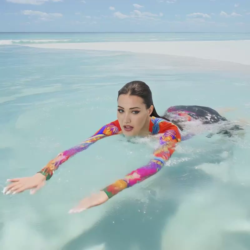 Close up angle front view of female swimming in the sea. She is wearing a a long-sleeved rash guard top with a vibrant, colorful tropical scene with insects. The top features a red base with blue, orange, purple, and yellow floral and abstract patterns.