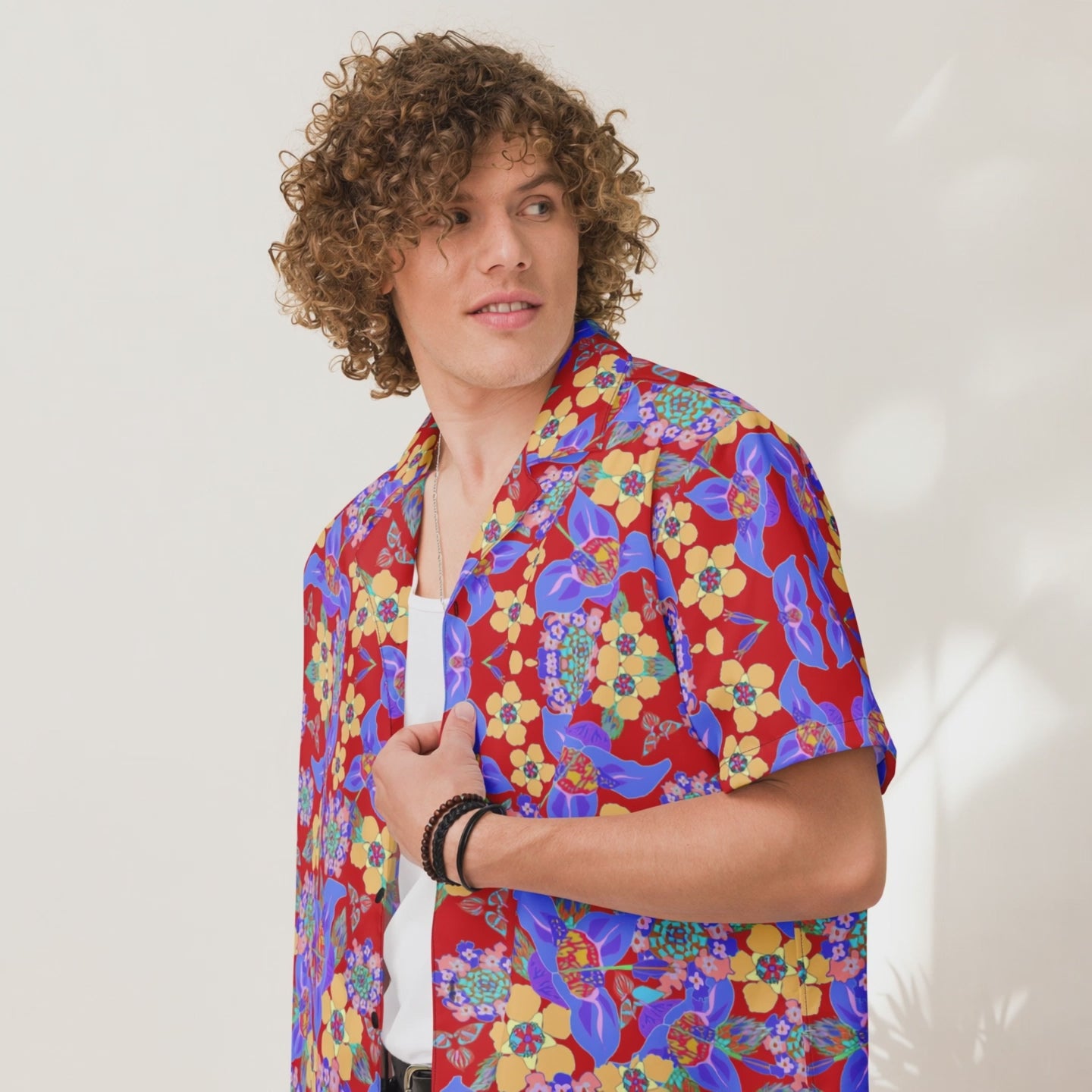 Curly-haired man wearing a red tropical floral oversized button shirt over a white tee, shown in a close-up side pose against a bright vacation-style backdrop.