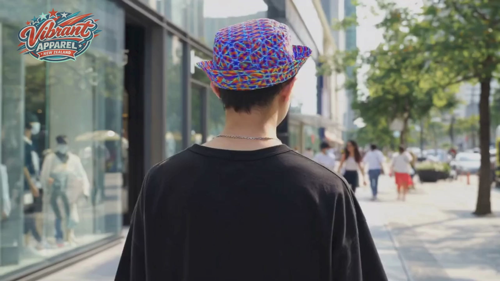 A cinematic clip of a young man walking down a busy city sidewalk, wearing a black oversized t-shirt and the "Vibrant Apparel" reversible bucket hat. The hat is styled on its blue and red kaleidoscopic side. As he walks, the camera orbits him, showing the hat from different angles against the backdrop of modern storefronts and city trees. The "Vibrant Apparel New Zealand" logo is visible in the top left corner.