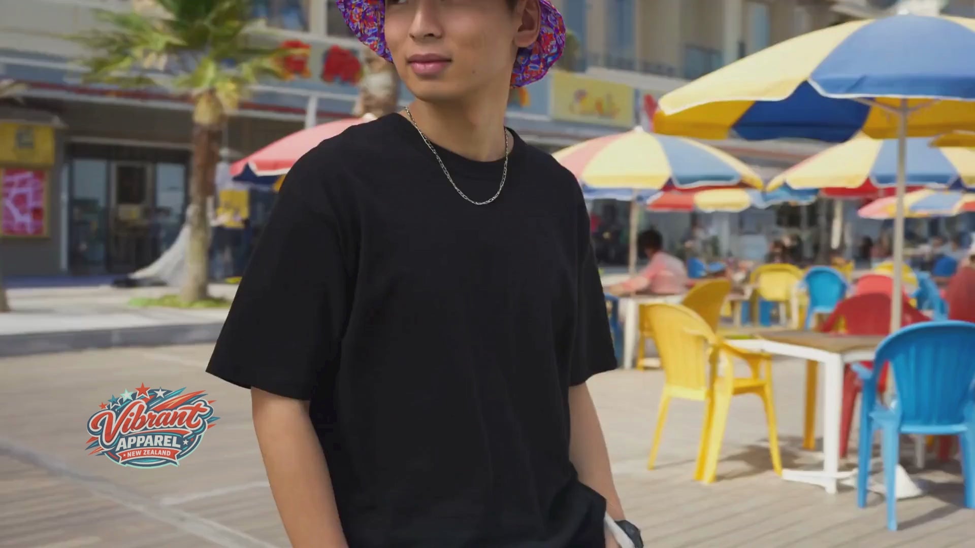 A vibrant video featuring the same young man, now on a sunny boardwalk with the ocean in the background. He is wearing the bucket hat on its purple and orange mandala side. The video shows him looking out at the waves and walking past colorful beach umbrellas, highlighting the hat as a versatile summer accessory. The high-energy edit features the "Vibrant Apparel" logo and a tropical, upbeat atmosphere.